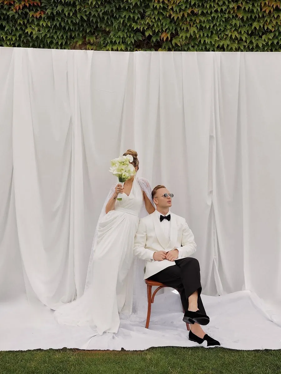 Novios frente al fondo del estudio montado en el jardín, ella de pie y él sentado